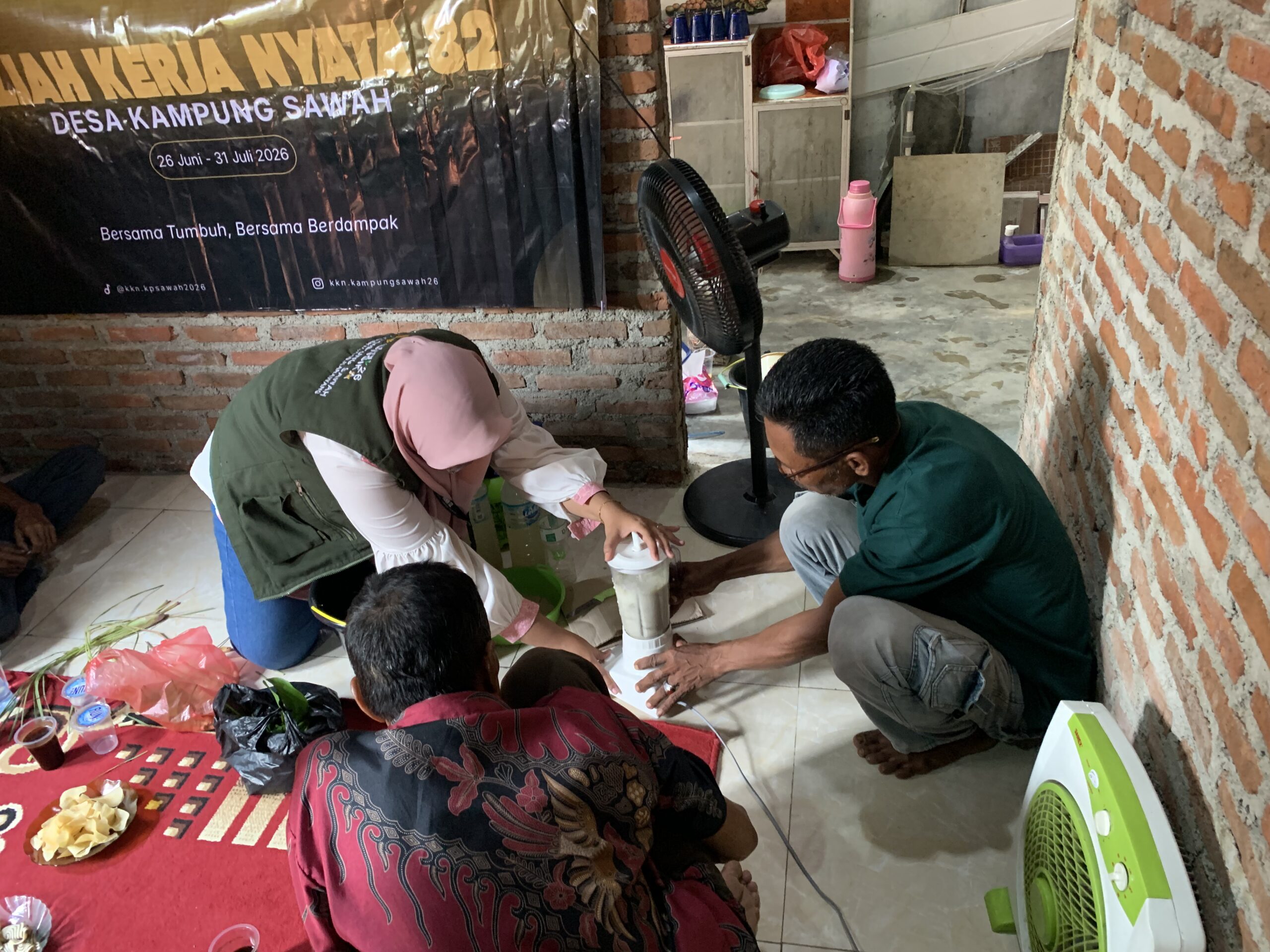 Dua orang petani dan seorang mahasiswa KKN sedang berjongkok di lantai, bersama-sama mengoperasikan blender yang berisi bahan berwarna putih, kemungkinan sedang dalam proses pembuatan pestisida nabati, di dekat spanduk KKN Desa Kampung Sawah.
