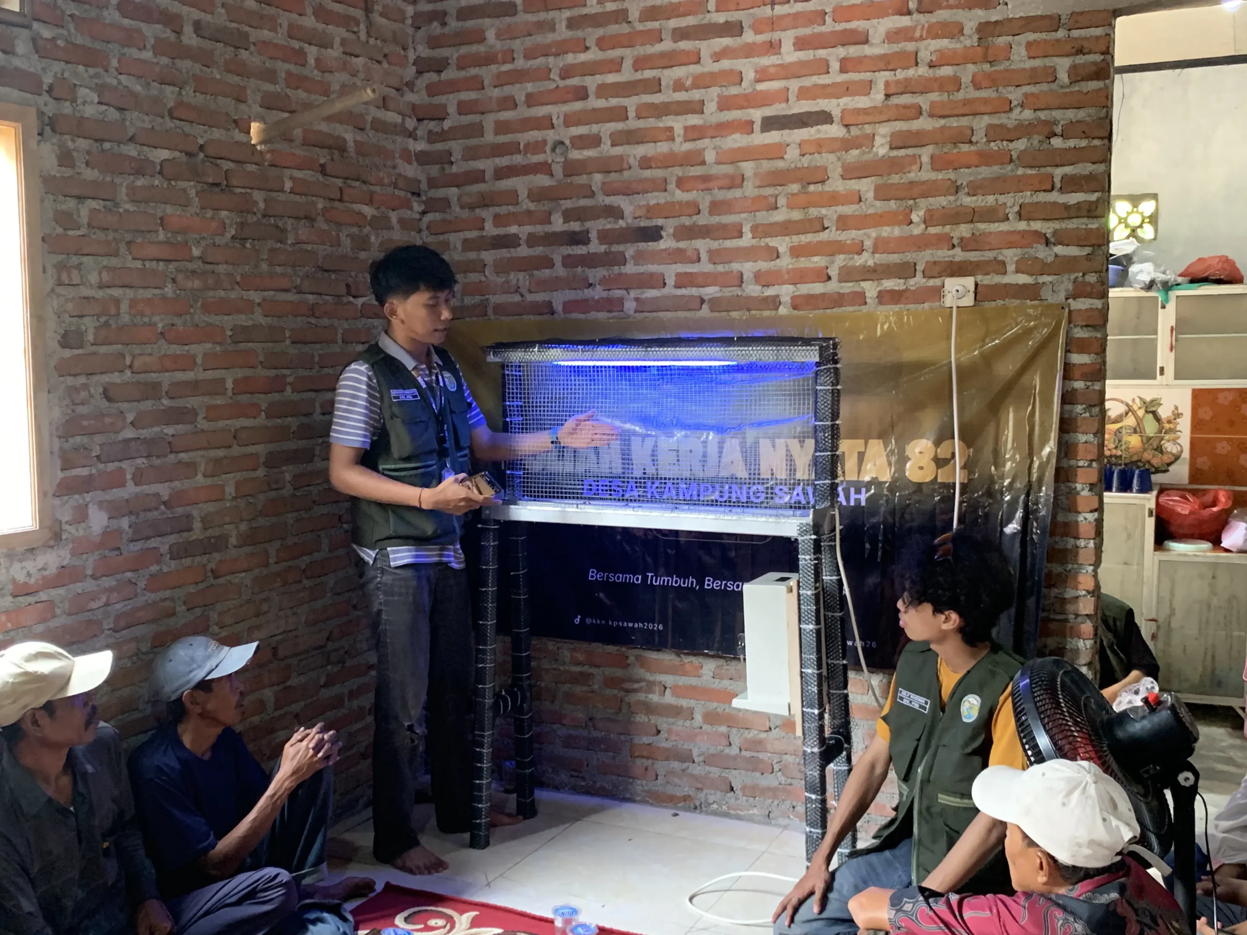 Seorang mahasiswa KKN berdiri di depan, memberikan penjelasan mengenai alat Net Electric Trap yang menyala dengan lampu biru kepada sekelompok petani desa yang duduk di lantai di dalam sebuah ruangan.